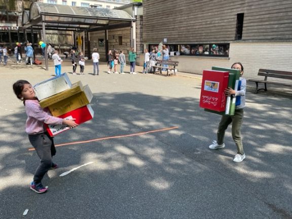 Unas alumnas cargando con libros gigantes de cartón