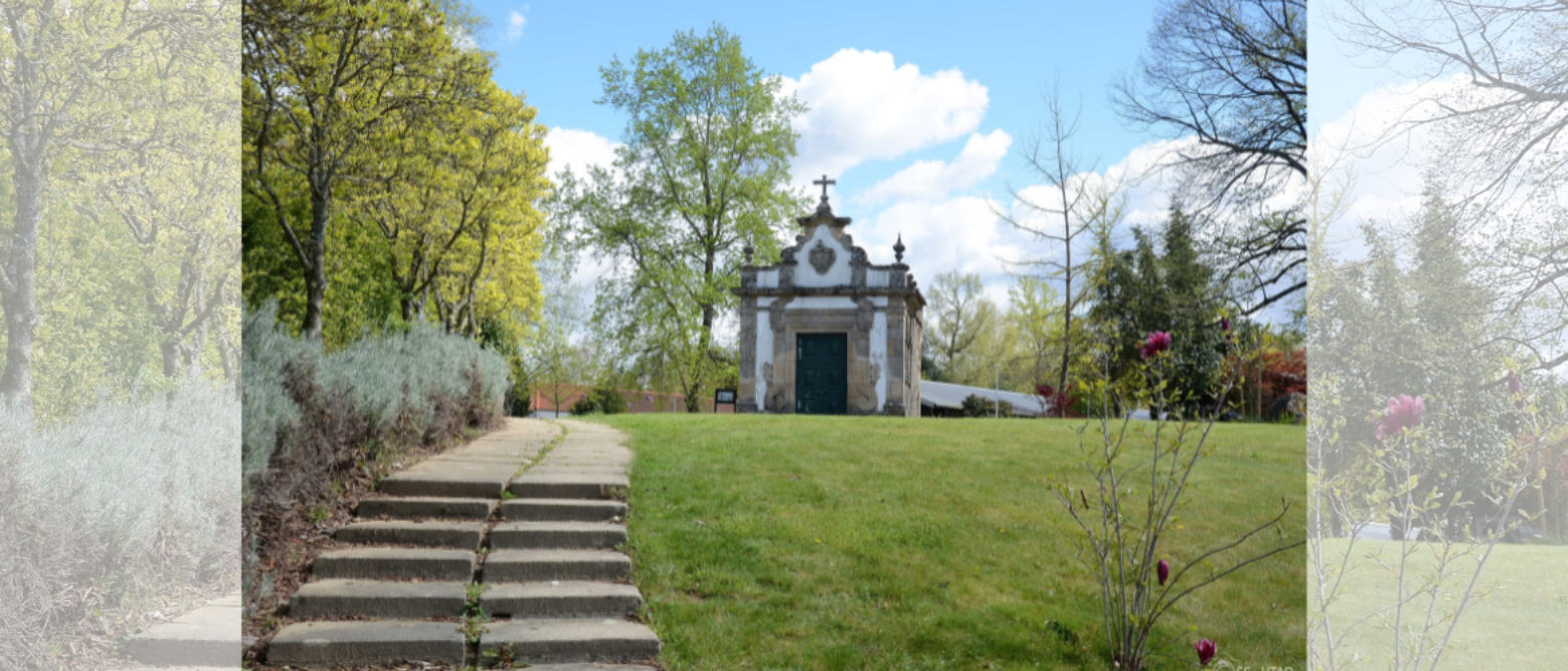 Pequeña capilla barroca blanca con cruz y ornamentos en piedra, rodeada de jardín con césped y árboles primaverales, accesible por escaleras de piedra