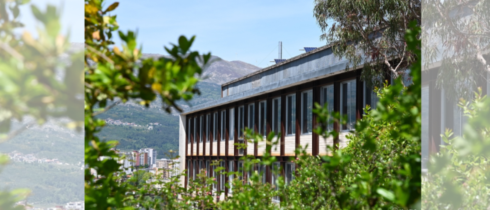 Edificio universitario moderno con grandes ventanales y estructura metálica, enmarcado por vegetación verde, con montañas y ciudad al fondo bajo cielo