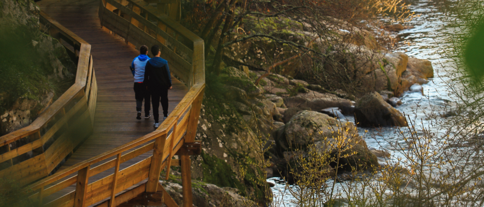 fotografía de dos personas paseando sobre un puente de madera mientras miran el paisaje lleno de árboles y rocas que dan a parar a un río