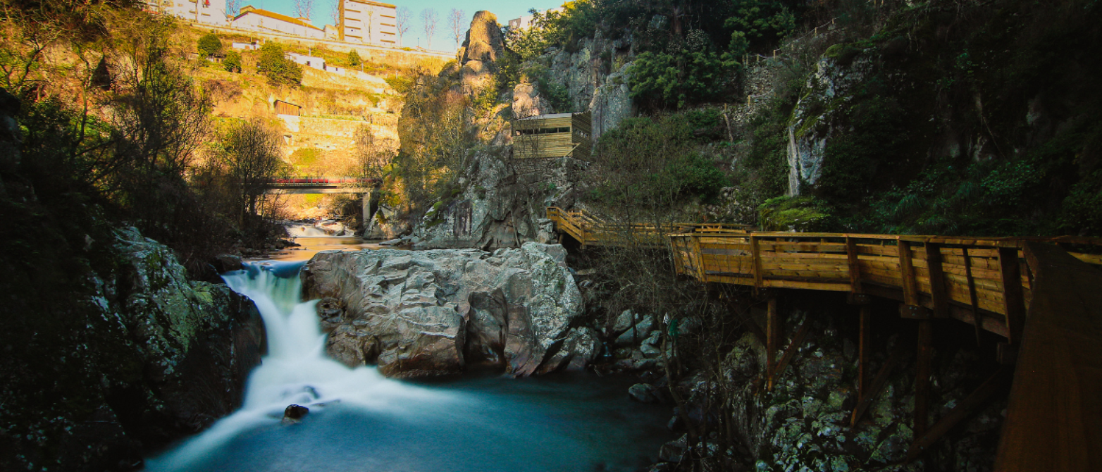 fotografía de un puente de madera mientras y el paisaje lleno de árboles y rocas que dan a parar a un río. Se ve un pequeña cascada con un alto caudal